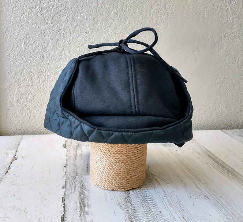 May include: A dark navy blue winter hat with a quilted brim and ear flaps. The hat has a soft, felt-like crown and a tie closure at the top. The hat is displayed on a cylindrical stand wrapped in natural twine.