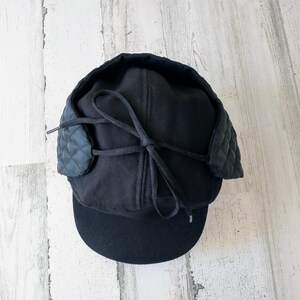 May include: Black winter hat with ear flaps. The hat features a quilted design on the ear flaps and a black brim. A black tie is used to secure the ear flaps. The hat is made of a soft, dark material.