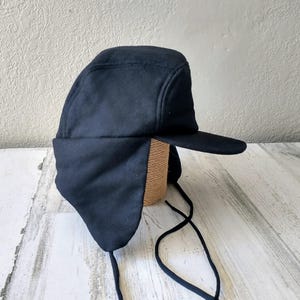 May include: A navy blue winter hat with ear flaps and a brim. The hat is made of a soft, dark fabric and has a structured crown. Black drawstrings hang from the ear flaps.