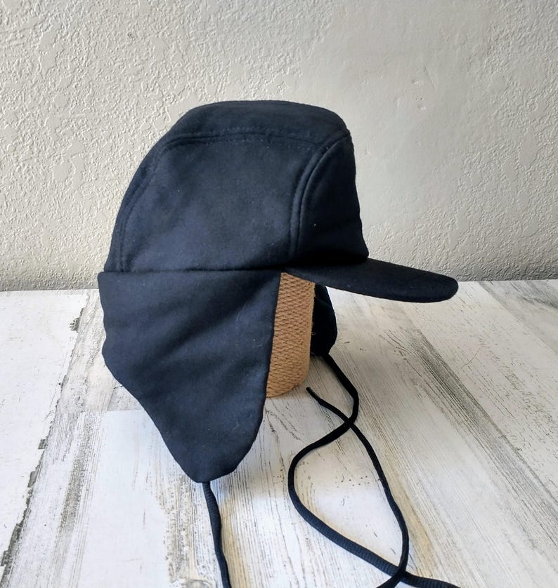 May include: A navy blue winter hat with ear flaps and a brim. The hat is made of a soft, dark fabric and has a structured crown. Black drawstrings hang from the ear flaps.