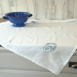 May include: White cotton tea towel with blue embroidered design of a cup and saucer. The towel is folded and lying on a white countertop.