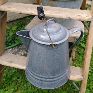 May include: A gray speckled enamel coffee pot with a conical body, a side handle, and a lid featuring a black wooden knob. The pot has a metal bail handle and shows signs of rust on the lid and near the spout, with a wide base and tapered top.
