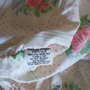 May include: Close-up of a vintage floral bed sheet. The sheet features a pattern of pink roses and green leaves on a white background with pink polka dots. The label reads "PERMA-PREST PERCALE".
