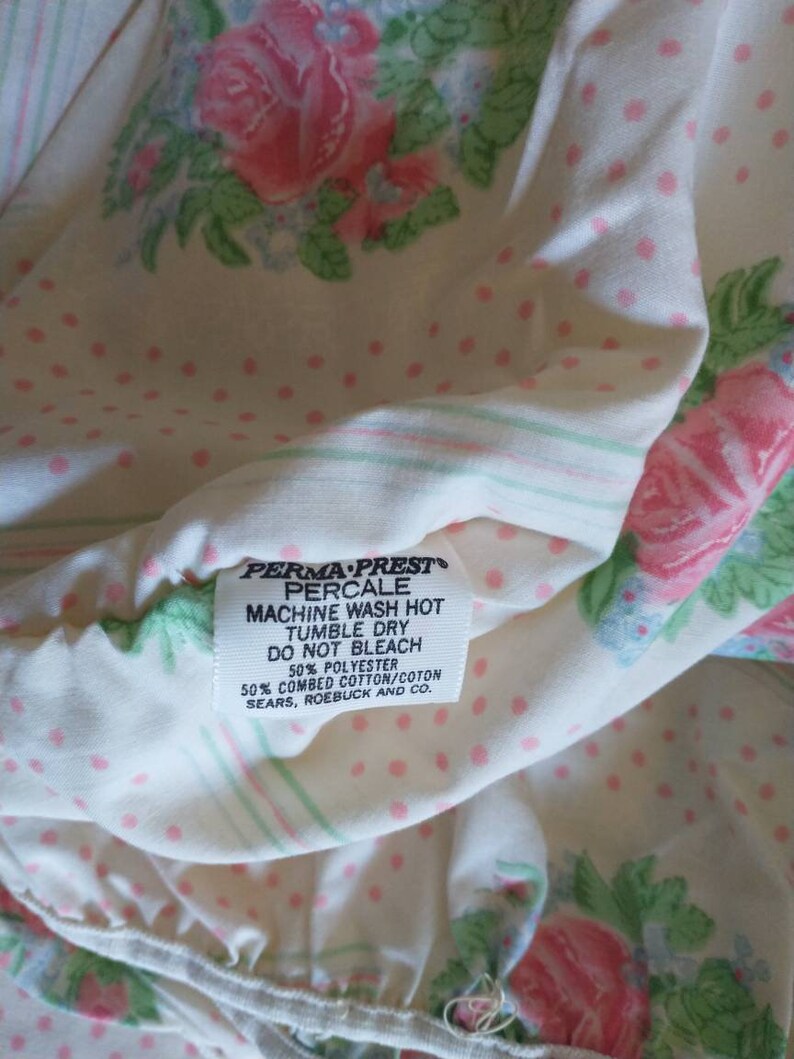 May include: Close-up of a vintage floral bed sheet. The sheet features a pattern of pink roses and green leaves on a white background with pink polka dots. The label reads "PERMA-PREST PERCALE".