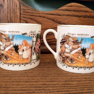 May include: Two white ceramic mugs with gold trim and a musical note design. The mugs feature a whimsical illustration of a group of people and the text "Allegro moderato".