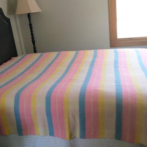 May include: A bed is covered with a pastel-striped blanket. The blanket features vertical stripes of pink, yellow, and blue against a white background. The bed is against a gray wall, with a lamp and window visible.