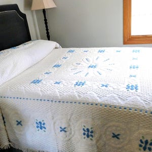 May include: A white bedspread with a floral pattern in light blue covers a bed. The bedspread has a textured surface and a fringe along the bottom edge. The headboard is dark gray.