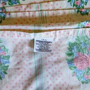 May include: Twin-size Perma-Prest percale sheet with a floral pattern. The sheet features pink roses, blue accents, and green leaves on a white background with pink polka dots. The label reads "PERMA-PREST PERCALE".