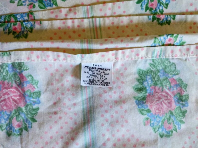 May include: Twin-size Perma-Prest percale sheet with a floral pattern. The sheet features pink roses, blue accents, and green leaves on a white background with pink polka dots. The label reads "PERMA-PREST PERCALE".