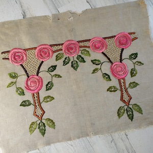May include: Embroidered floral design featuring pink roses and green leaves on a beige fabric. The embroidery has a detailed, textured appearance, with a brown outline and a beige mesh-like center. The fabric has a distressed, vintage look.
