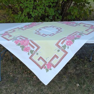 May include: A white tablecloth with a yellow center and a brown and yellow geometric pattern. The tablecloth has a pink floral design around the border.