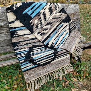 May include: A woven rug with a geometric pattern in shades of black, white, brown, and blue. The rug has fringe on all sides and is folded in half.
