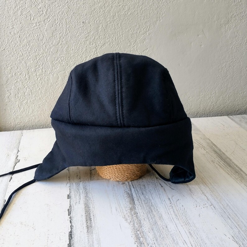 May include: A dark navy blue winter hat with ear flaps and a stitched seam down the center. The hat has long black ties and is made of a soft, warm material. This hat is perfect for cold weather.