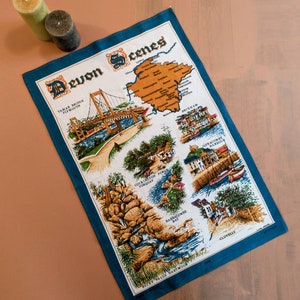 May include: A blue tea towel with a white background featuring a map of Devon, England, with illustrations of various landmarks and the text "Devon Scenes".