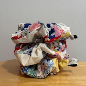 May include: A colorful scrunchie made from patchwork fabric, featuring a variety of patterns and colors including red, blue, yellow, and white. The scrunchie is made of multiple fabric pieces sewn together. A small tag with the word "EVELYN" is attached.