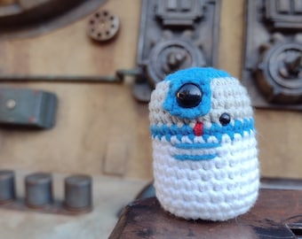 May include: A crocheted plush toy resembling a droid from the Star Wars franchise. The toy is white with blue and gray accents and has a black eye.