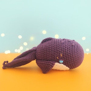 May include: A purple crocheted whale plush toy with a white belly and blue eyes. The whale is lying on its side on an orange surface.