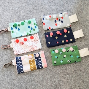 Puede incluir: Seis bolsas de tela con diferentes patrones y colores. Cada bolsa tiene una correa blanca y una fila de botones coloridos. Las bolsas son perfectas para guardar objetos pequeños como llaves, bálsamo labial o auriculares.