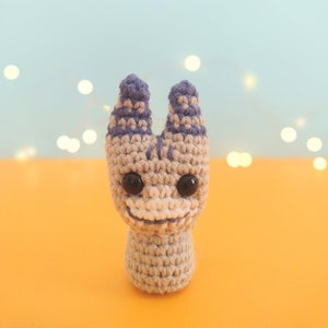 May include: A handmade crochet creature with a smiling face, large black eyes, and two upright ears. The crochet toy is primarily beige with purple accents and is set against a yellow and blue background.