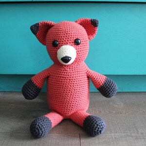 Puede incluir: Un juguete de peluche de zorro de crochet con un cuerpo rojo, orejas y pies negros, y nariz y boca blancas.