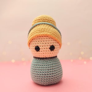 Puede incluir: Una muñeca de crochet con un sombrero amarillo y un vestido azul. La muñeca tiene ojos negros y una expresión neutral.