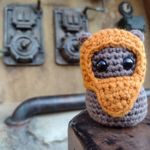 May include: A small crocheted plush toy with an orange helmet and brown body. The toy has large black eyes and a small mouth.