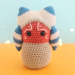 Peut inclure: Un jouet en peluche crocheté avec un visage rouge, un corps gris et un couvre-chef rayé bleu et blanc. Le jouet a des yeux noirs et une bouche cousue.