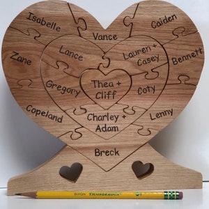 May include: A wooden heart-shaped puzzle with names inscribed on each piece. The names include Isabelle, Lance, Zane, Vance, Thea + Cliff, Gregory, Copeland, Caiden, Lauren + Casey, Coty, Charley + Adam, Lenny, and Bennett. The puzzle is on a wooden stand with two heart-shaped cutouts.