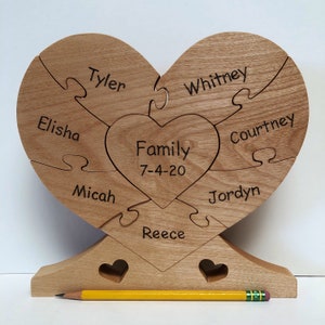 May include: A wooden heart-shaped puzzle with the names Tyler, Elisha, Micah, Reece, Whitney, Courtney, Jordyn, and the text "Family 7-4-20" engraved on it. The puzzle is assembled and standing on a wooden base.