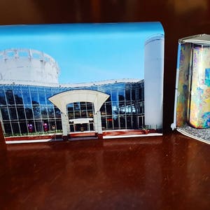 May include: A miniature diorama of a building with a glass facade and a dome-shaped roof. The diorama is open to reveal a small interior scene with two miniature figures, a clock, and a colorful patterned cylinder.
