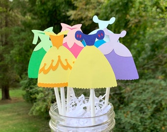 May include: Six paper princess dress cupcake toppers in a glass jar. The toppers are in various colors, including green, yellow, pink, and purple. The dresses are decorated with details like ruffles and bows.