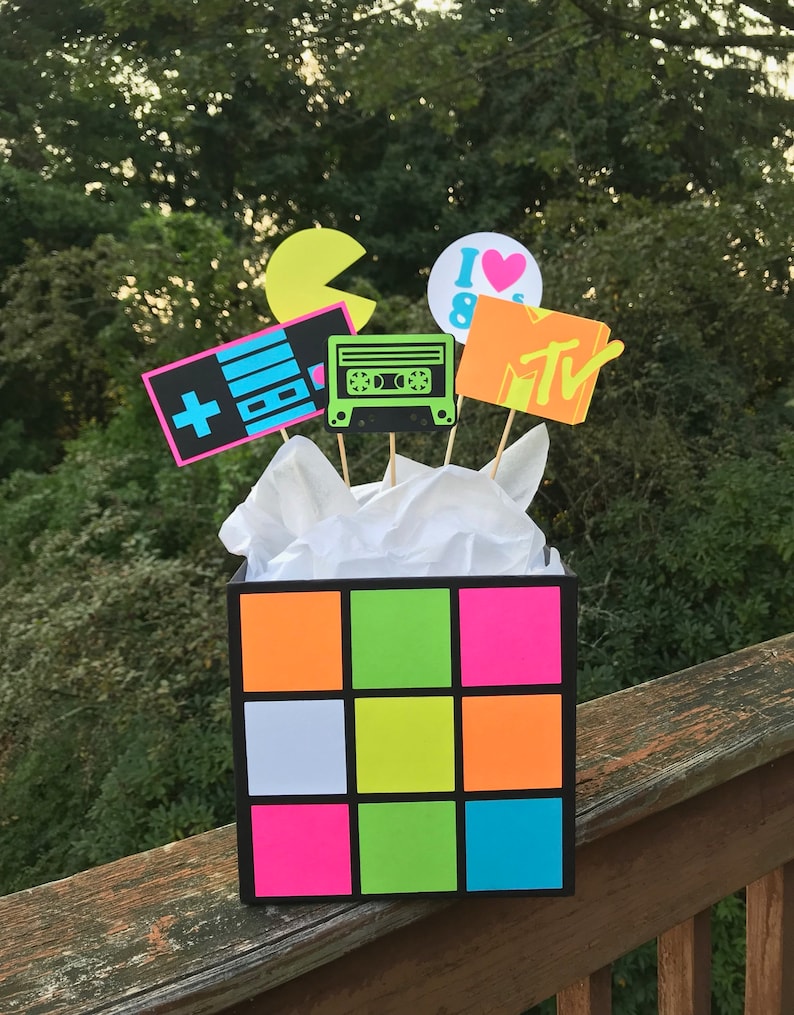 May include: A black box with a Rubik's Cube design, filled with 1980s-themed cutouts on sticks. These include a Pac-Man, a cassette tape, a video game controller, an MTV logo, and an "I ❤️ 80s" graphic. The box is filled with white tissue paper.
