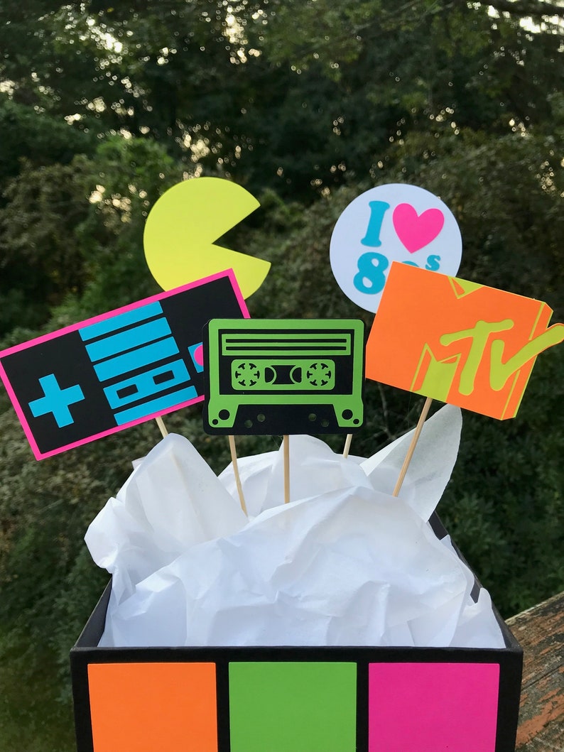 May include: A black box with neon orange, green, and pink panels. The box contains paper and several 80s-themed cutouts on sticks, including a Pac-Man, a cassette tape, a controller, an MTV logo, and an "I ❤️ 80s" design.
