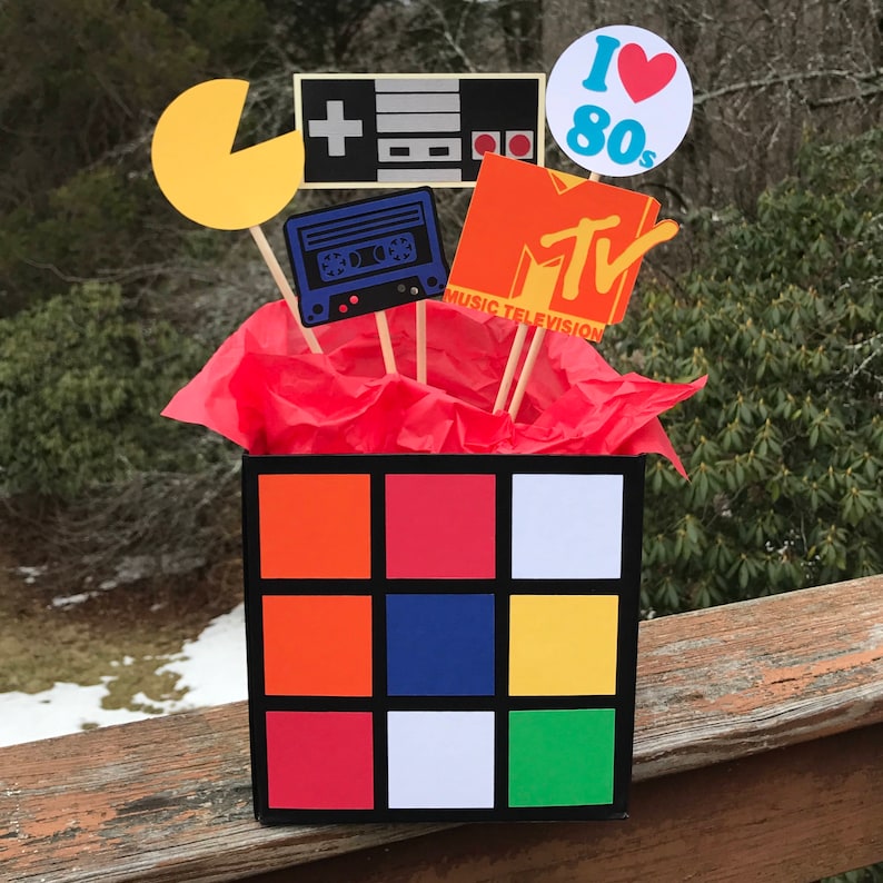May include: A retro-themed centrepiece with a Rubik's Cube box filled with red tissue paper. It's topped with cutouts of a Pac-Man, a video game controller, a cassette tape, an MTV logo, and an "I Love 80s" circle.