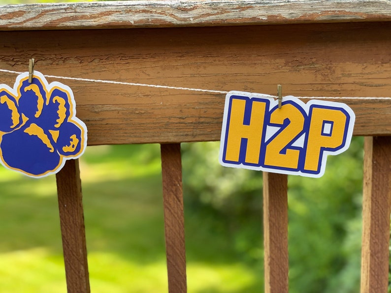 May include: A white string with a blue paw print and a yellow banner with the text "H2P" hanging from a wooden railing.
