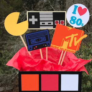 May include: A vibrant 80s-themed display with cutouts of a Pac-Man, a games console, a cassette tape, and the MTV logo. The display includes the text "I Love 80s". The cutouts are on wooden sticks, arranged in a black box with orange, red, and white squares.