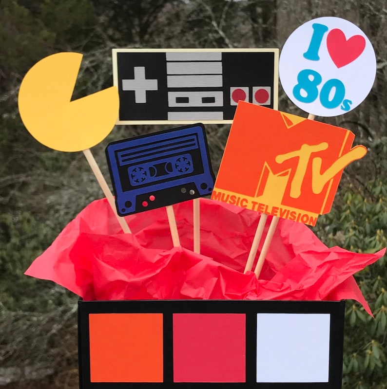 May include: A vibrant 80s-themed display with cutouts of a Pac-Man, a games console, a cassette tape, and the MTV logo. The display includes the text "I Love 80s". The cutouts are on wooden sticks, arranged in a black box with orange, red, and white squares.