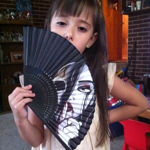 A little girl holding a folding hand fan that reveals the art on it. The art shows a pale skinned woman, crying black tears.