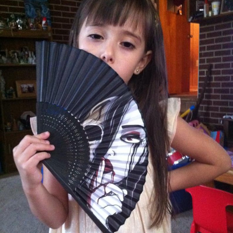 A little girl holding a folding hand fan that reveals the art on it. The art shows a pale skinned woman, crying black tears.