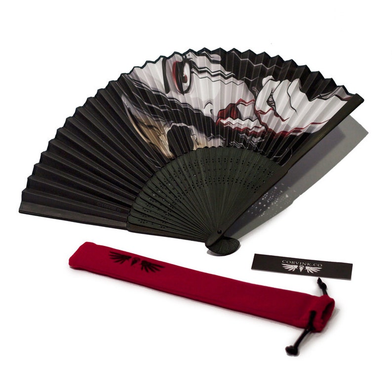 A black folding hand fan made of paper and bamboo. The paper part of the fan has an illustration of a colorless skinned woman, who has a skull for half her face. She is shushing and dripping blood from her mouth.