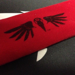 Black crow skull logo with wings screen printed on a red fabric pouch.