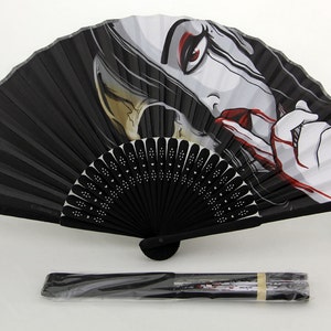 A fully extended black hand fan that has an illustration of a woman who resembles death. Half of her face is a skull, she is bleeding from her mouth and crying black tears. There is also a folded and sealed fan in the foreground.