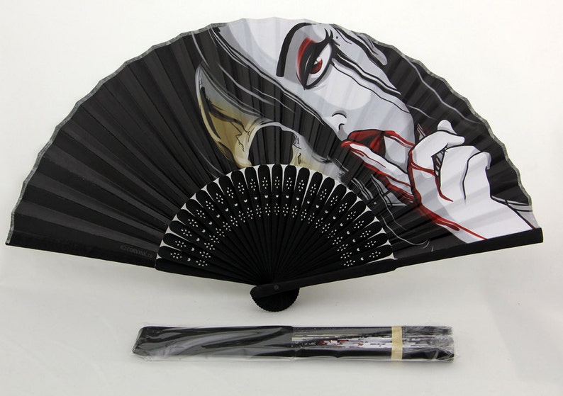 A fully extended black hand fan that has an illustration of a woman who resembles death. Half of her face is a skull, she is bleeding from her mouth and crying black tears. There is also a folded and sealed fan in the foreground.