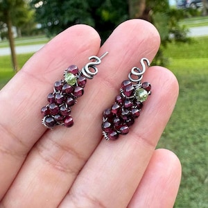 May include: A pair of silver-toned earrings shaped like grape clusters. Each earring features a cluster of small, dark red beads with a single green bead and a decorative silver wire design.