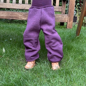 Może przedstawiać: Fioletowe spodnie z polaru z wysokim stanem i ciemniejszymi fioletowymi mankietami. Spodnie wykonane są z miękkiego, teksturowanego materiału. Zdjęcie przedstawia spodnie na dziecku stojącym na zielonej trawie.