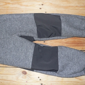 Może przedstawiać: Szare spodnie dresowe z czarnymi łatkami na kolanach. Spodnie dresowe mają gruby pas i ściągacze na kostkach.