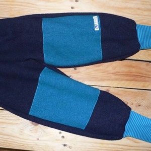 Könnte beinhalten: Blaue und türkisfarbene gestreifte Strickhose mit kontrastierendem dunkelblauem Stoff. Die Hose hat ein Etikett mit der Aufschrift "Little Lambs" auf der Vorderseite.