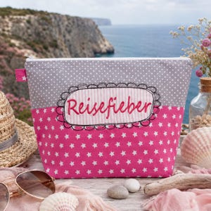 Puede incluir: Un estuche con cremallera que presenta una sección superior gris con lunares blancos y una sección inferior rosa con estrellas blancas. Un parche ovalado central con borde negro festoneado contiene el texto rosa bordado "Reisefieber" sobre fondo a rayas.