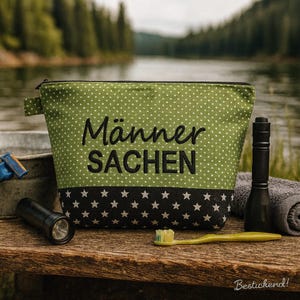 May include: A green wash bag with white polka dots and the words "Männer Sachen" in black. The bottom of the bag has a black band with white stars. A black torch, a toothbrush, and a towel are next to the bag.