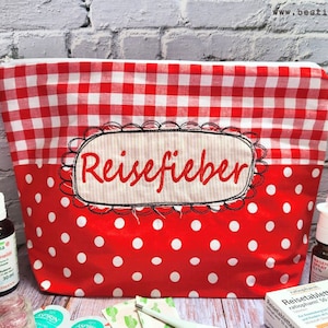 Op de afbeelding: Rode en witte gingham en stippenstof met rits met het woord "Reisefieber" geborduurd op de voorkant. De tas is gevuld met verschillende medicijnen en reisbenodigdheden in reisformaat.